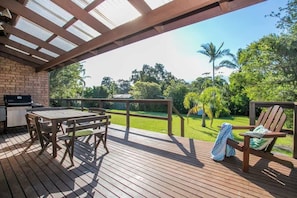 An outdoor patio with a wooden table and chairs, a grill, and a view of a lush garden. - Family Paradise at Mollymook (Mollymook)