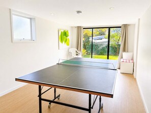 Un tavolo da ping pong in una stanza con una grande finestra, una sedia bianca e un tavolino con un cesto.