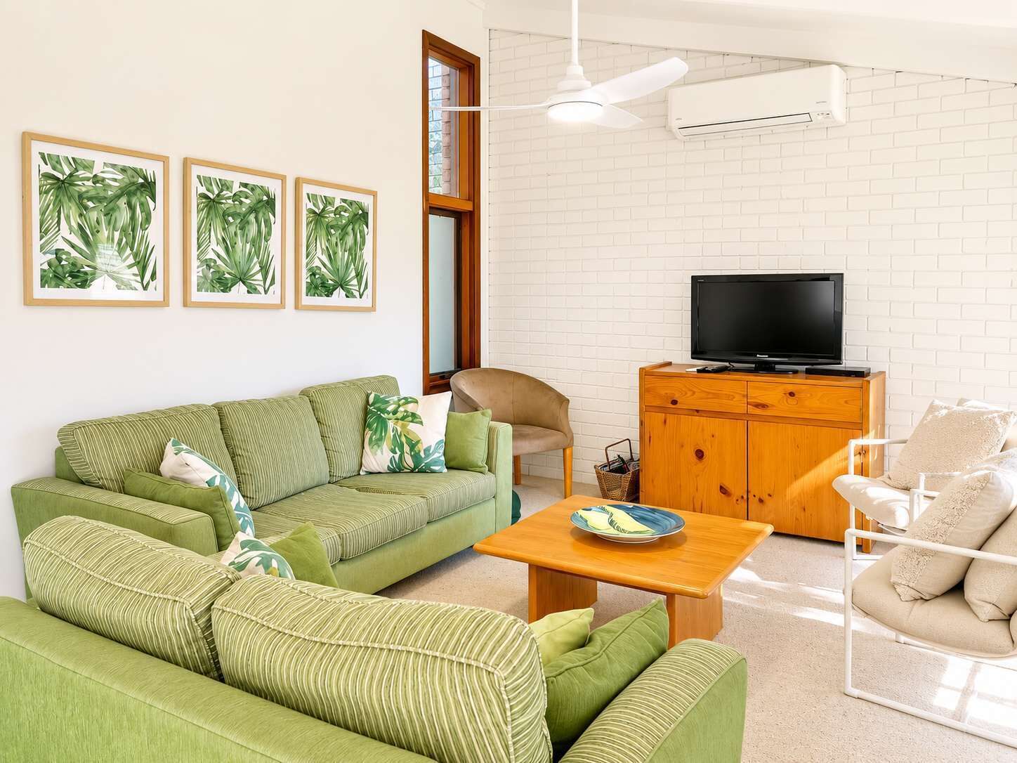 Una sala de estar con un sofá verde, un mueble de madera con un televisor y obras de arte con hojas tropicales enmarcadas en la pared.