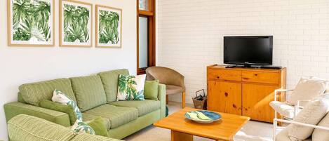 Una sala de estar con un sofá verde, un mueble de madera con un televisor y obras de arte con hojas tropicales enmarcadas en la pared.