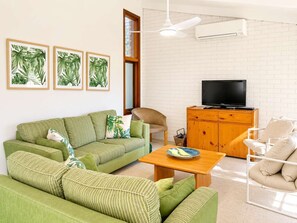 A living room with a green sofa, a wooden cabinet with a TV, and framed tropical leaf artwork on the wall. - Family Paradise at Mollymook (Mollymook)