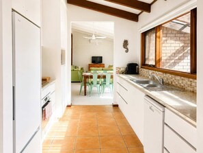 A kitchen with a tiled floor, a dining area with chairs, a television, and a window with a view of a brick wall. - Family Paradise at Mollymook (Mollymook)