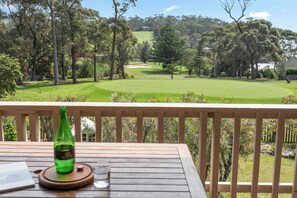 A wooden deck with a bottle and glass, overlooking a golf course with trees and a distant building. - Tee to Sea - 3BR home with golf & ocean views (Mollymook)