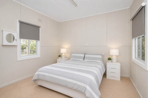 A bedroom with a bed, two bedside tables with lamps, a window with blinds, and a wall-mounted air conditioning unit. - So Close to the Beach (Mollymook)
