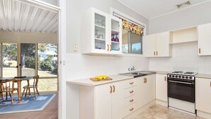 A kitchen with white cabinets, a stove, and a dining area with chairs and a table. - So Close to the Beach (Mollymook)