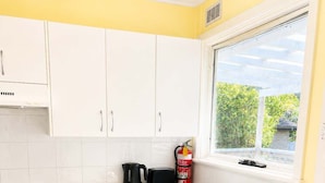 Cocina compacta con armarios blancos, estufa a gas, cafetera y extintor cerca de la ventana.