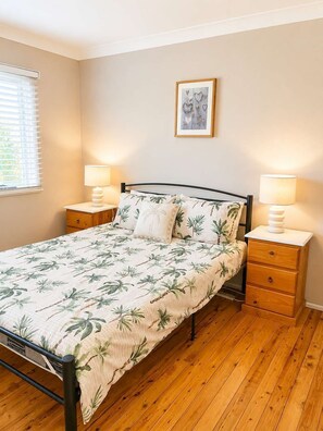Une chambre à coucher comprenant un lit, deux tables de chevet, une commode, une fenêtre avec des stores et un tableau accroché au mur.