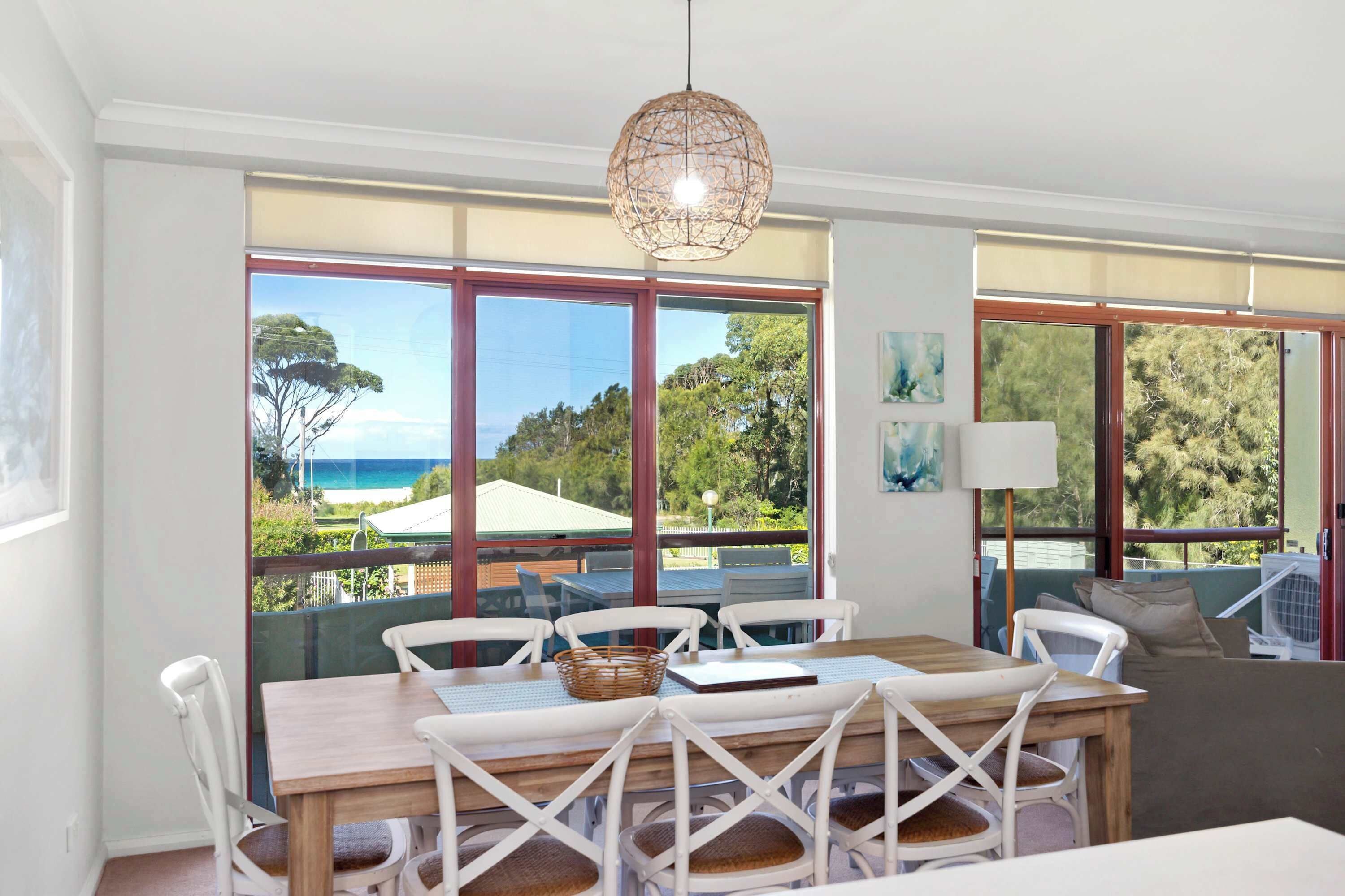 A dining area with a wooden table and white chairs, large windows with a view of the sea, and a hanging light fixture.
