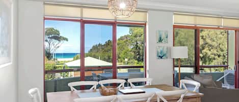 A dining area with a wooden table and white chairs, large windows with a view of the sea, and a hanging light fixture.