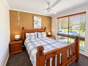 Un dormitorio con cama de madera, ventilador de techo, mesita de noche con lámpara, una obra de arte en la pared y una ventana con persianas.