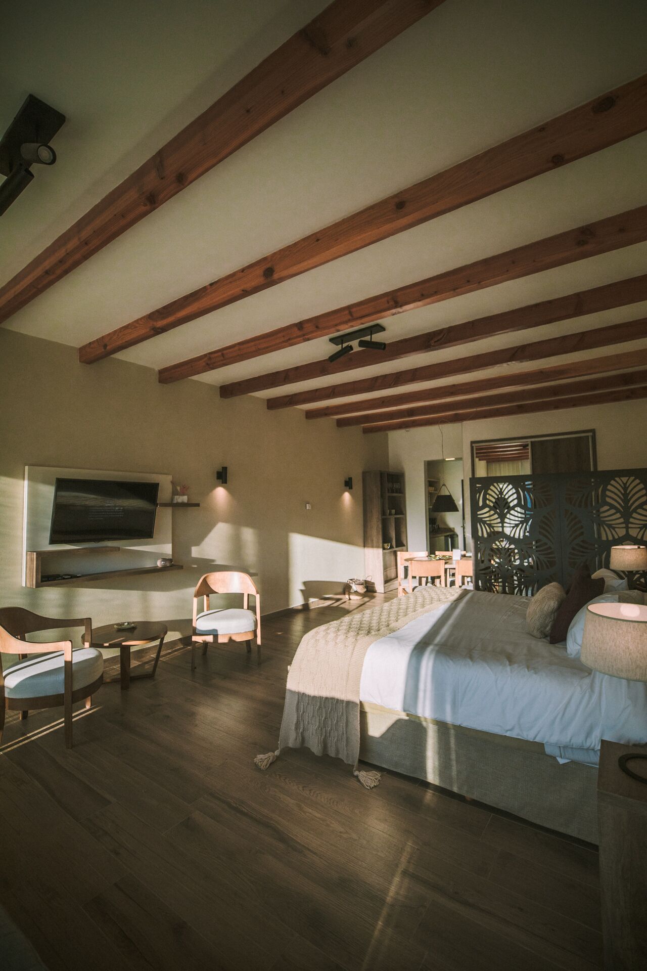 A spacious bedroom with a large bed, wooden beams, a flat-screen TV, and a seating area with chairs and a table.