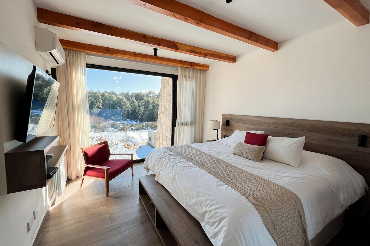 A hotel room with a large bed, a television, a red chair, and a view of a snowy landscape through the window.