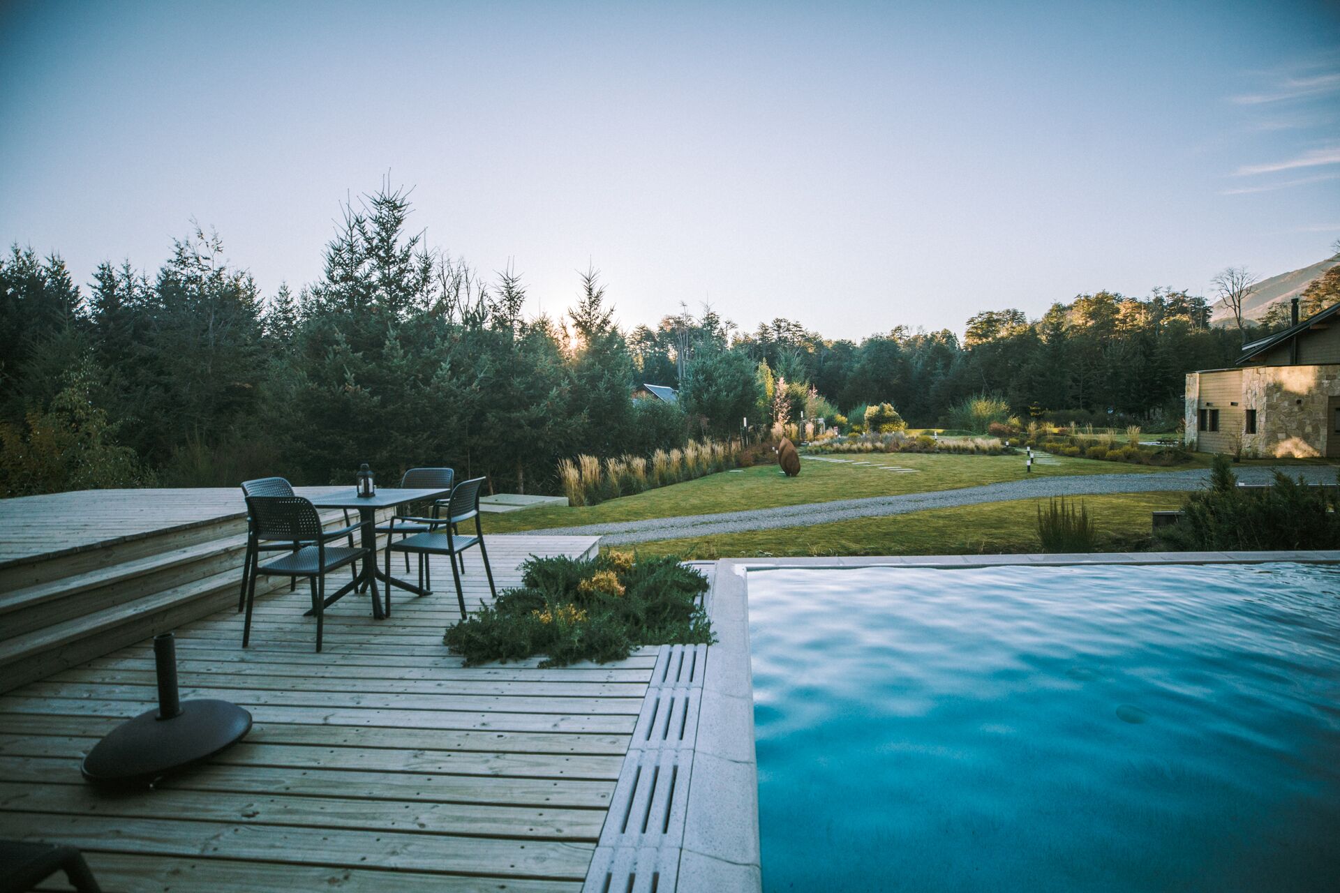 A wooden deck with outdoor furniture, a swimming pool, and a landscaped garden with a view of a house and trees.