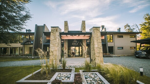 A modern hotel entrance with stone pillars, a gravel driveway, and a well-maintained garden with various plants and a seating area. - Awka Apart Hotel (Villa La Angostura)