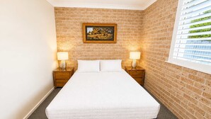 Une chambre d’hôtel avec un mur en briques, un lit avec une literie blanche, deux tables de chevet avec des lampes et une fenêtre avec des stores.