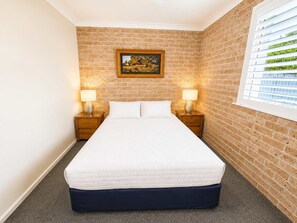 A hotel room with a brick wall, a bed with white bedding, two bedside tables with lamps, and a window with blinds. - Magnificent Outlook (Mollymook Beach)
