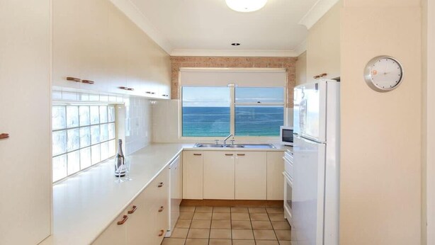 Une cuisine compacte avec vue sur la mer, des meubles blancs, un évier et une horloge au mur.