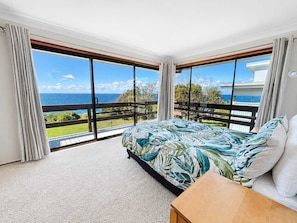 A bedroom with a large bed, a wooden nightstand, and a view of the ocean through floor-to-ceiling windows.