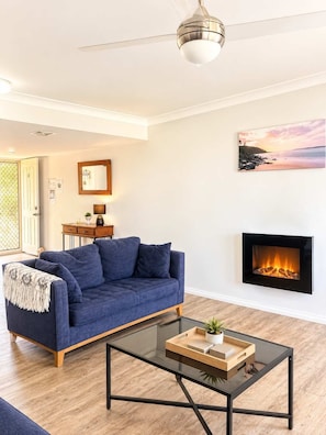 Una sala de estar con un sofá azul, una mesa de centro de vidrio, una chimenea y un ventilador de techo.