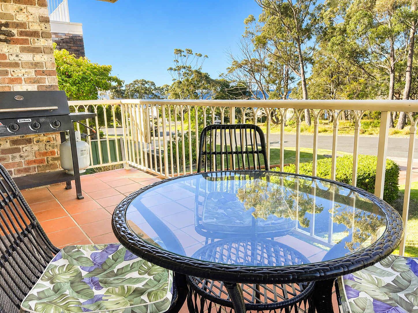 A glass-top table with chairs on a balcony, a barbecue grill, and a view of the surrounding greenery.