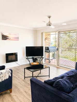 A living room with a flat-screen TV, a fireplace, a ceiling fan, and a sliding glass door leading to an outdoor grill area. - Perfect Location Near Collers Beach (Mollymook)
