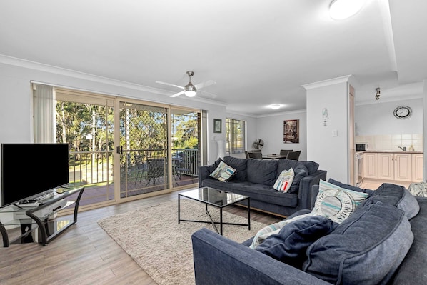 Un salon comprenant un canapé, une table basse, un téléviseur et un balcon avec vue sur les arbres.