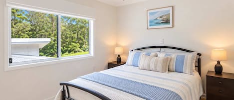 A bedroom with a bed, two bedside tables, a ceiling fan, a window with a view of greenery, and a framed picture on the wall.