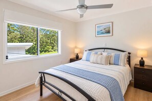 A bedroom with a bed, two bedside tables, a ceiling fan, a window with a view of greenery, and a framed picture on the wall.