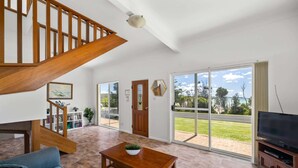 A spacious living room with a wooden staircase, a television, and a large sliding glass door leading to a patio. - Seaside Beach House (Dolphin Point)