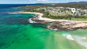 An aerial view of a coastal area with clear turquoise waters, a sandy beach, and a residential area with houses. - Seaside Beach House (Dolphin Point)