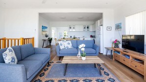 A living room with a sofa, coffee table, TV stand, and a view into the kitchen. - Seaside Beach House (Dolphin Point)
