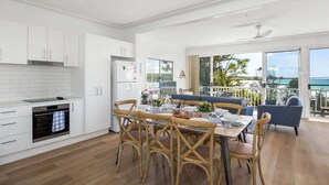 A modern kitchen with white cabinets, a dining area with wooden chairs, and a balcony with a view of the sea. - Seaside Beach House (Dolphin Point)