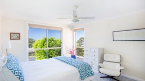 Relax in this serene bedroom with a large bed, ceiling fan, and a window offering a view of lush greenery.