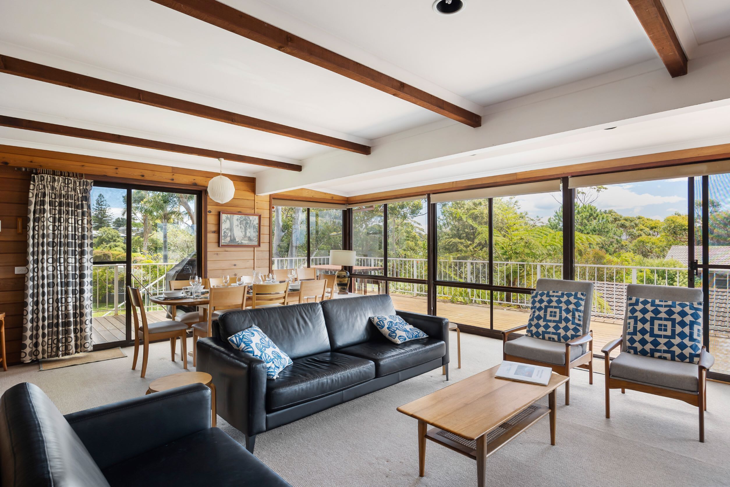 A modern living room with a leather sofa, wooden coffee table, and patterned armchairs. Large windows offer a view of the outdoors.