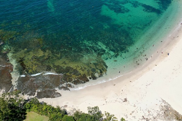 An aerial view of a beach with clear water, sandy shore, and rocky areas.