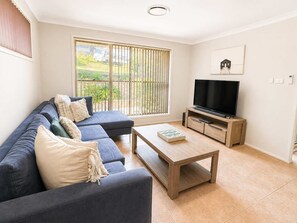A living room with a sofa set, a coffee table, a TV on a wooden stand, and a window with blinds. - Leafy Retreat at Ulladulla (Ulladulla)