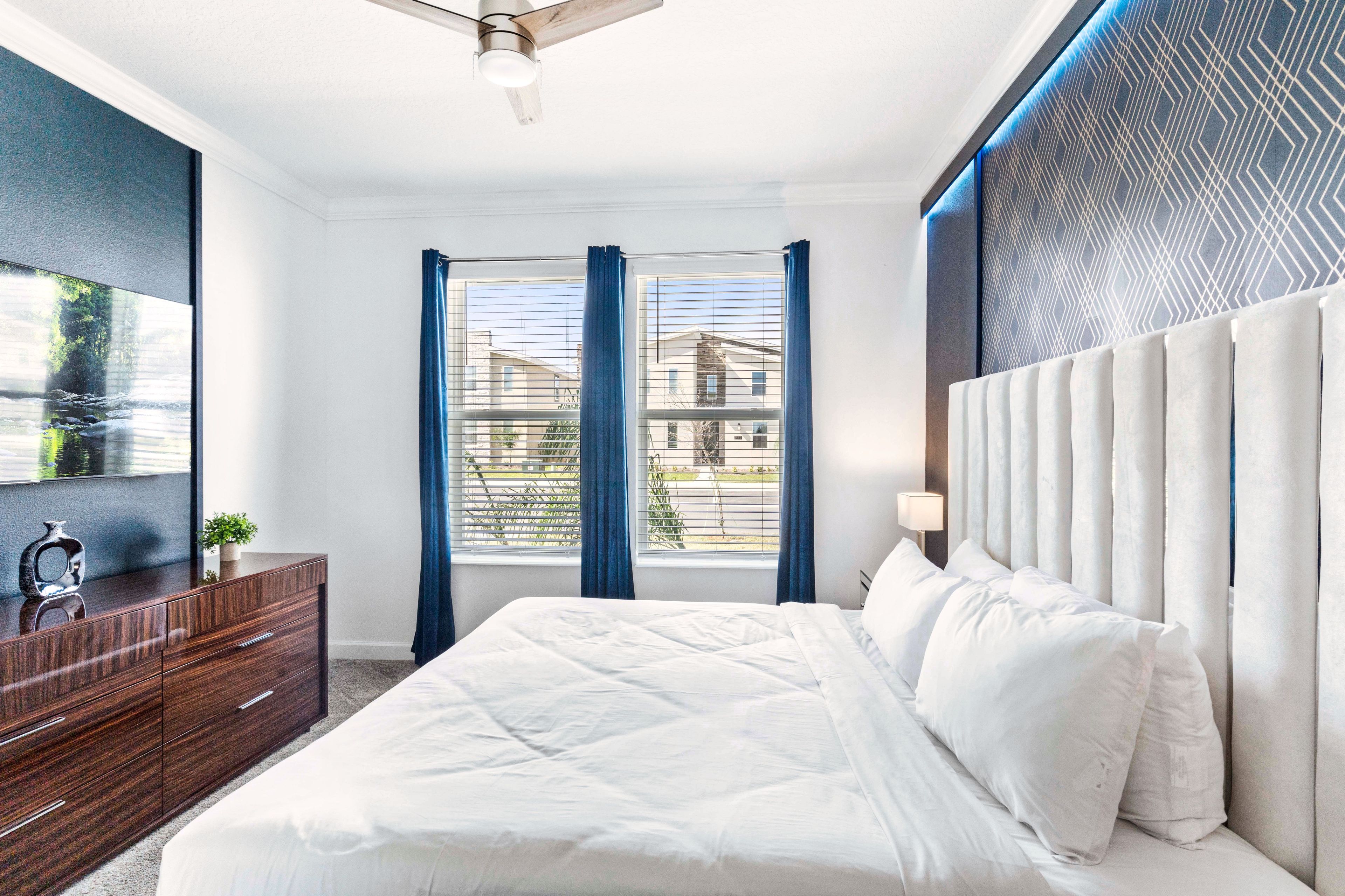 A modern bedroom with a large bed, a dresser, a ceiling fan, and a window with curtains.