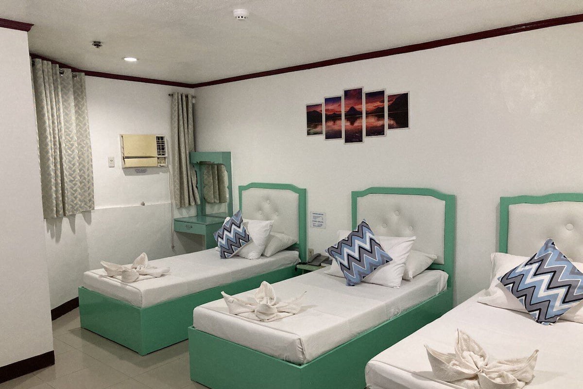 A hotel room with two single beds, each with a green frame and white bedding. There is a small bedside table with a lamp, a mirror, and a window with curtains.