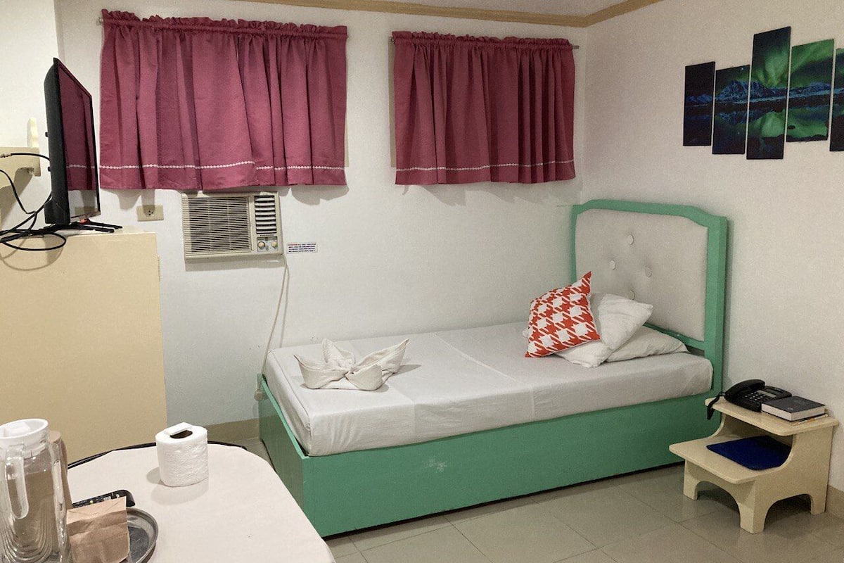 A small, single-bed room with a green headboard, a bedside table with a phone and books, a window with purple curtains, and a wall-mounted air conditioning unit.