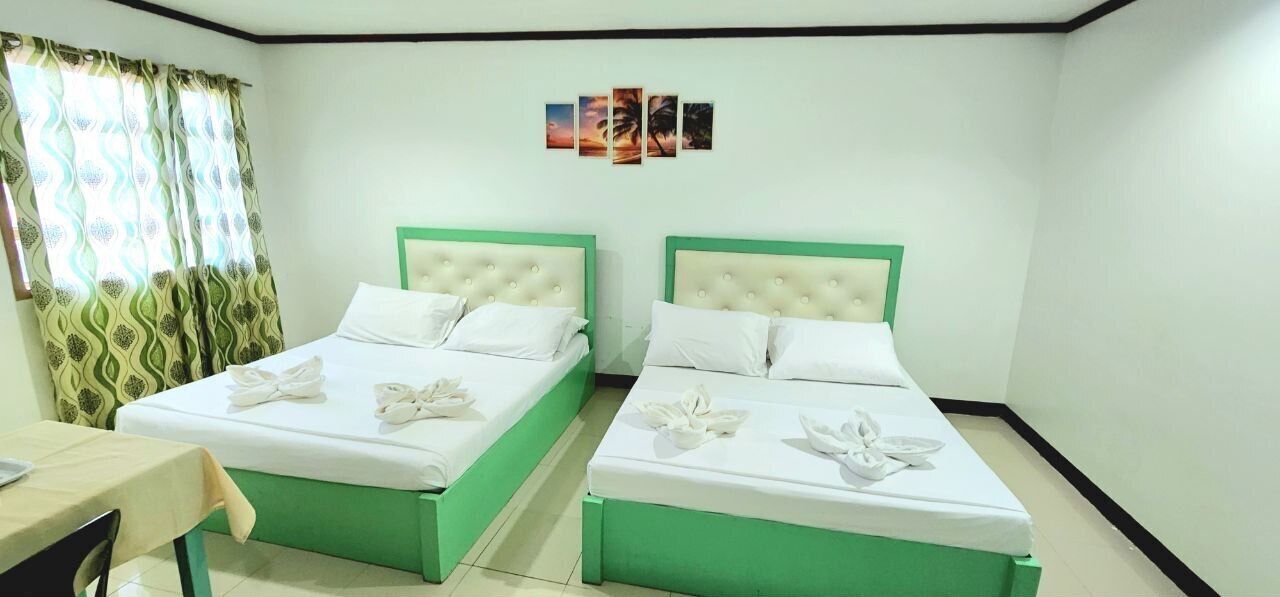 Ein Hotelzimmer mit zwei Betten, beide mit grünem Gestell und weißer Bettwäsche. Ein kleiner Tisch mit einem Stuhl steht dort, sowie ein Fenster mit gemusterten Vorhängen.