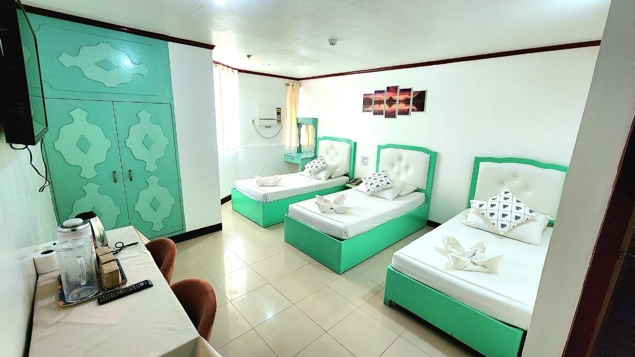 A hotel room with three beds, a desk with a computer and a coffee maker, and a green wardrobe.
