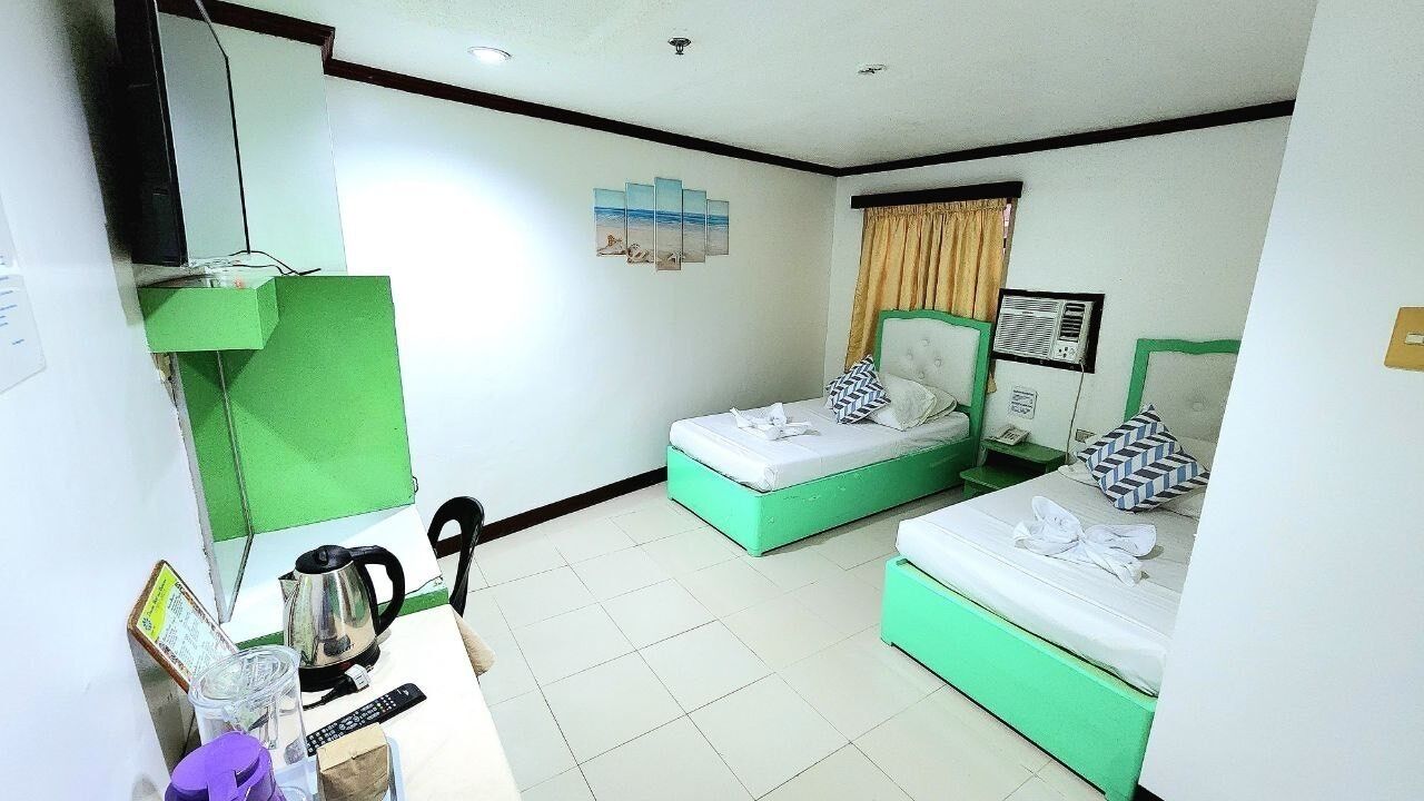 A hotel room with two single beds, a desk with a kettle, a chair, and a TV.