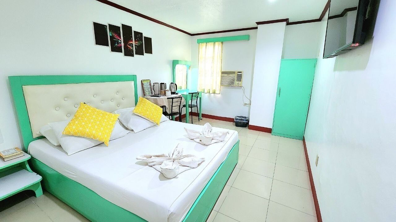 Quarto de hotel com cama, escrivaninha, cadeira, televisão e guarda-roupa.