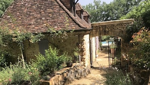 A rustic stone house with a brick roof, surrounded by a garden with potted plants and a stone pathway.