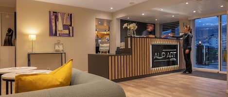 A hotel reception area with a modern design, a reception desk, a sofa with a yellow pillow, and a large window offering a view.