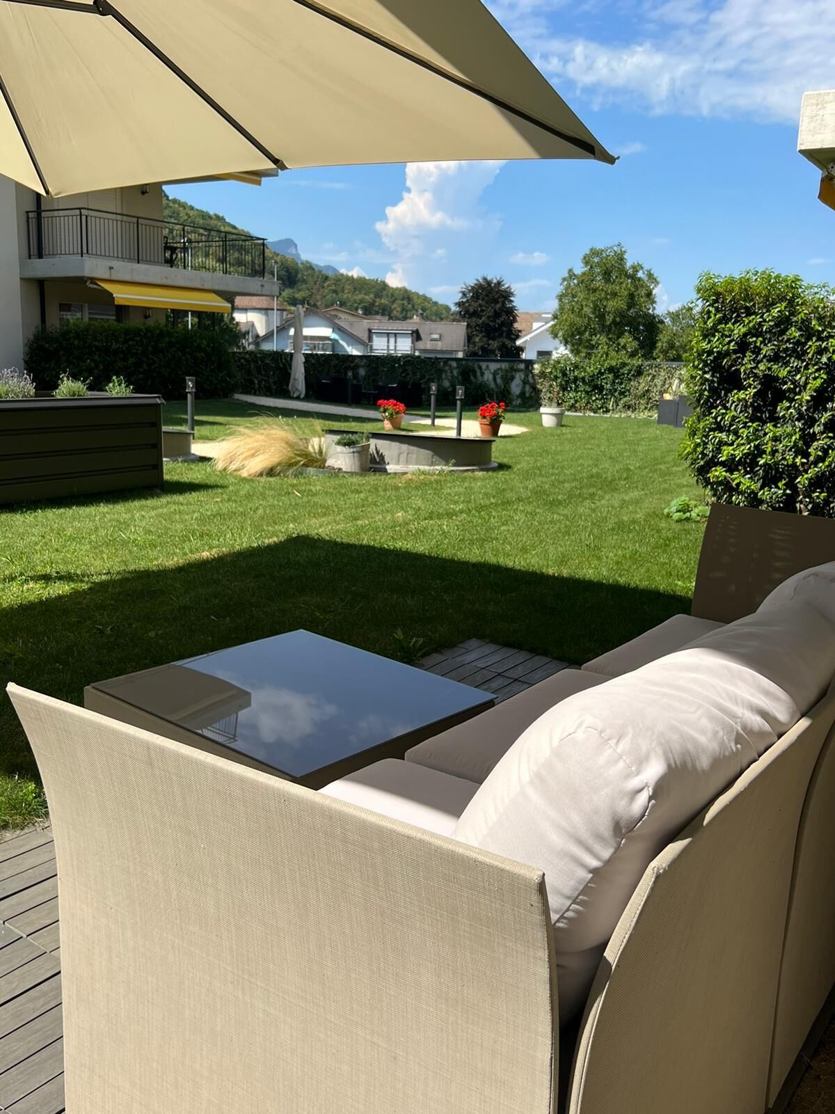 Een terras met een comfortabele bank en een tafel met glazen blad, een grote parasol en uitzicht op een verzorgd gazon en omliggende huizen.