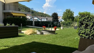 Een terras met een comfortabele bank en een tafel met glazen blad, een grote parasol en uitzicht op een verzorgd gazon en omliggende huizen.