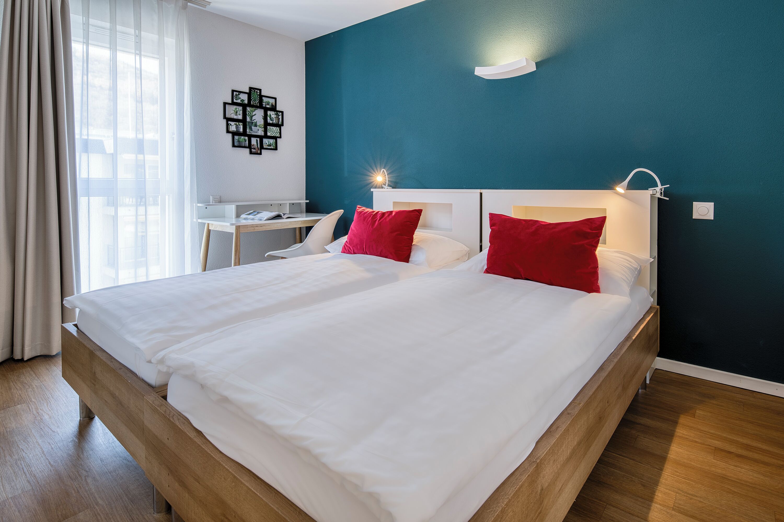 A double bed with white bedding and red pillows, a desk with a chair, a wall-mounted light, and a teal accent wall.