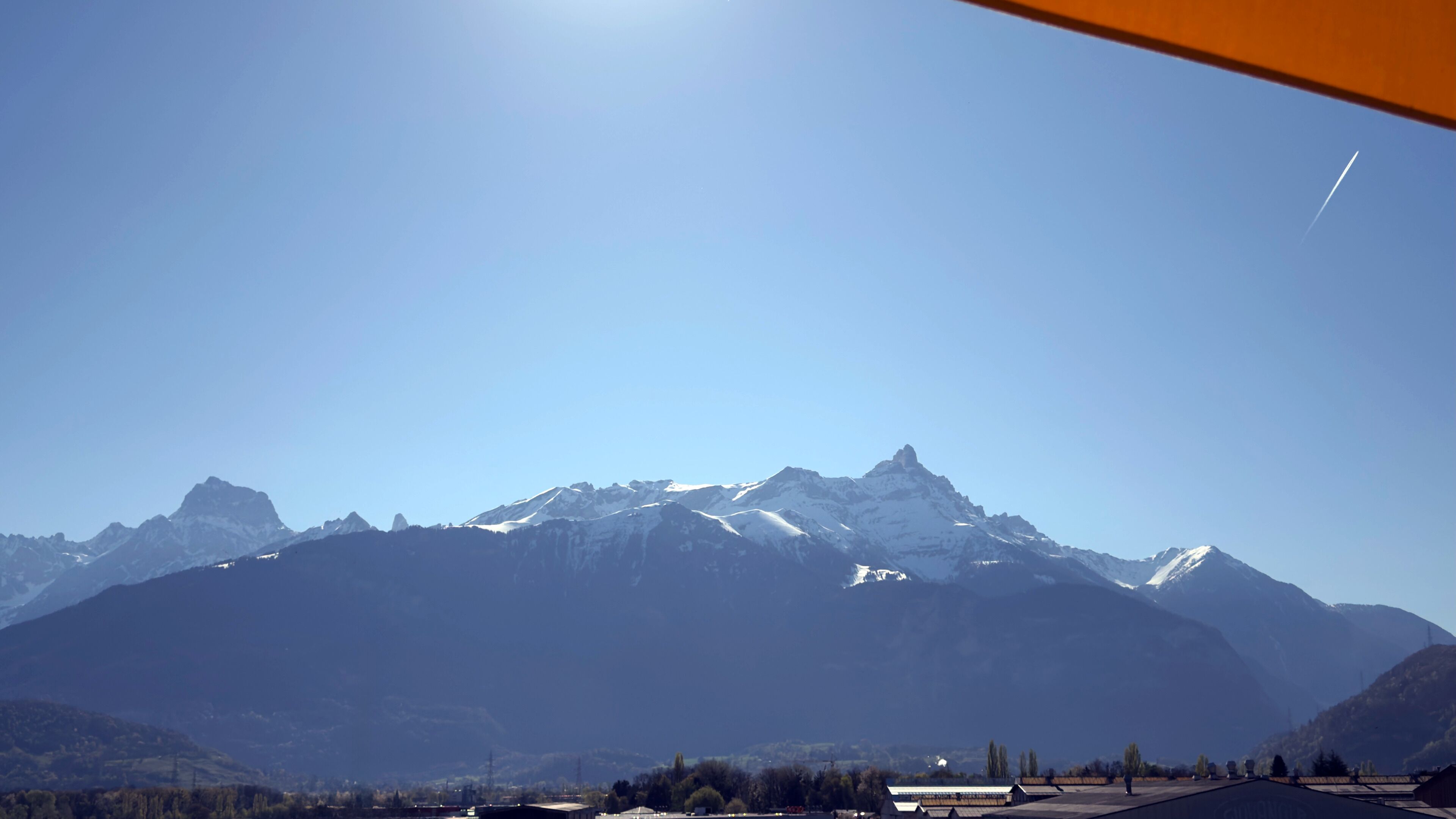 A mountain range with snow-capped peaks, a clear sky, and a distant view of a town with buildings and trees.