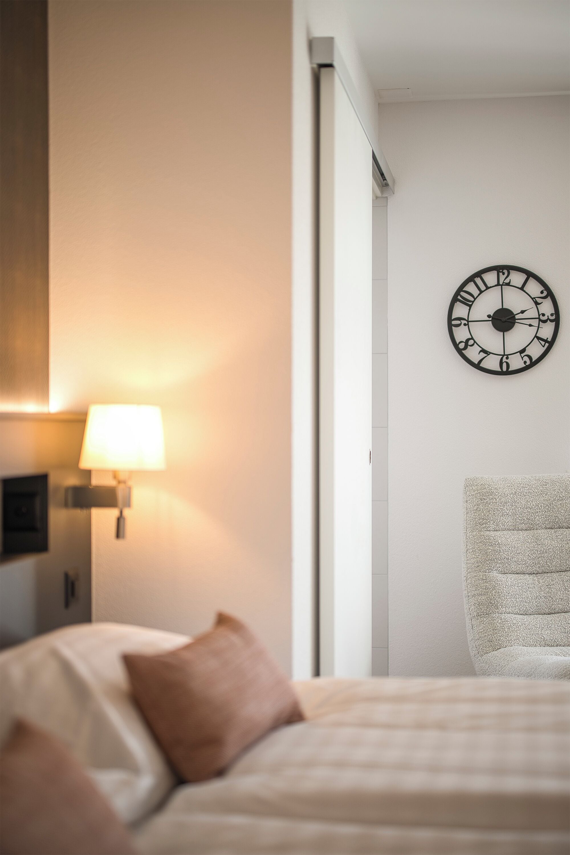 Una habitación de hotel con una cama, un reloj en la pared, una lámpara y una silla.